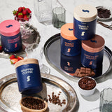 Assorted containers of whipped coffee, vanilla crème, peanut butter, and chocolate soufflé on a decorative tray with strawberries and chocolate.